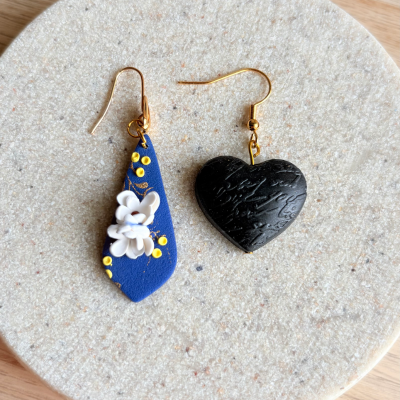 Orecchini artigianali in argilla polimerica con fiore bianco su base blu e cuore nero inciso. Gioielli simbolici fatti a mano da Daiana Aretes Studio